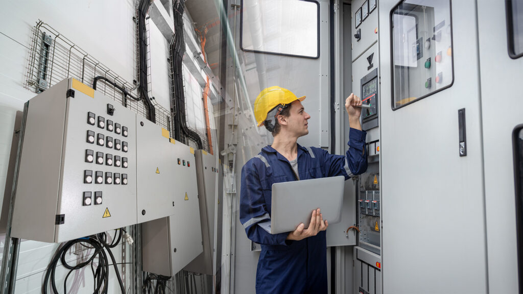 Technician wearing a safety helmet inspects an industrial control panel, using a laptop and digital tools to manage an IIoT project on Proficloud.io.