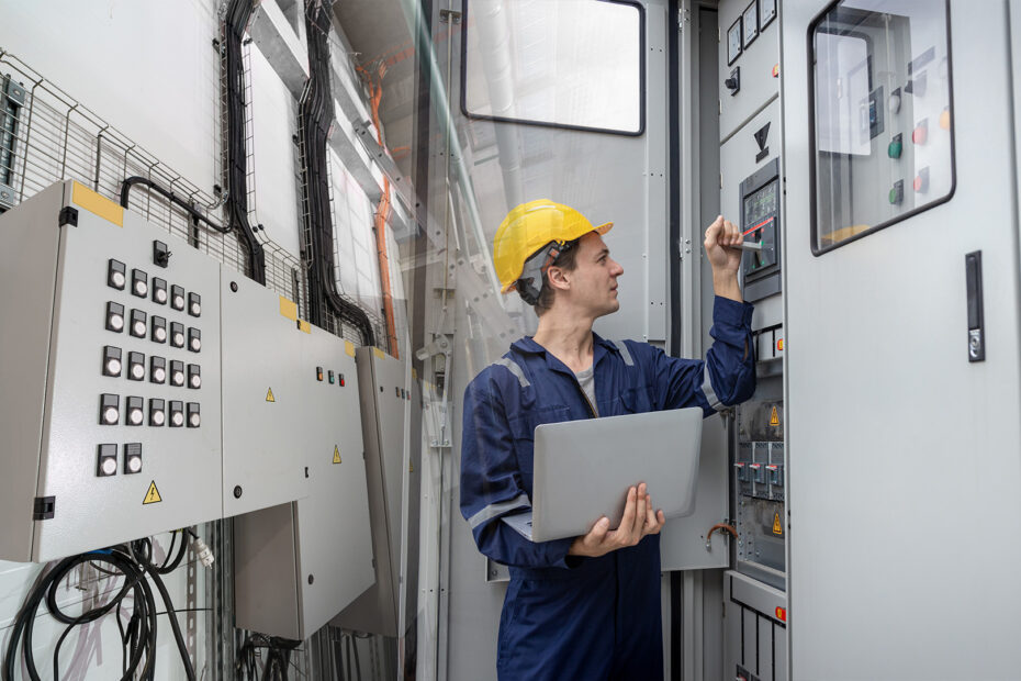 Technician wearing a safety helmet inspects an industrial control panel, using a laptop and digital tools to manage an IIoT project on Proficloud.io.